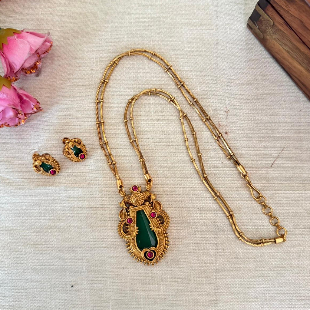 Gold necklace with green pendant and earrings on a textured surface with pink flowers.